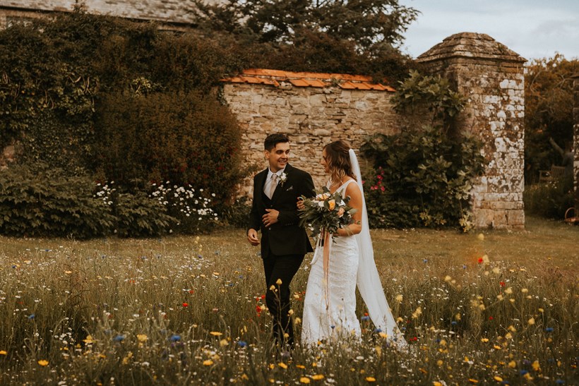 Bride and groom walk through wildflower meadow Ash Barton wedding venue