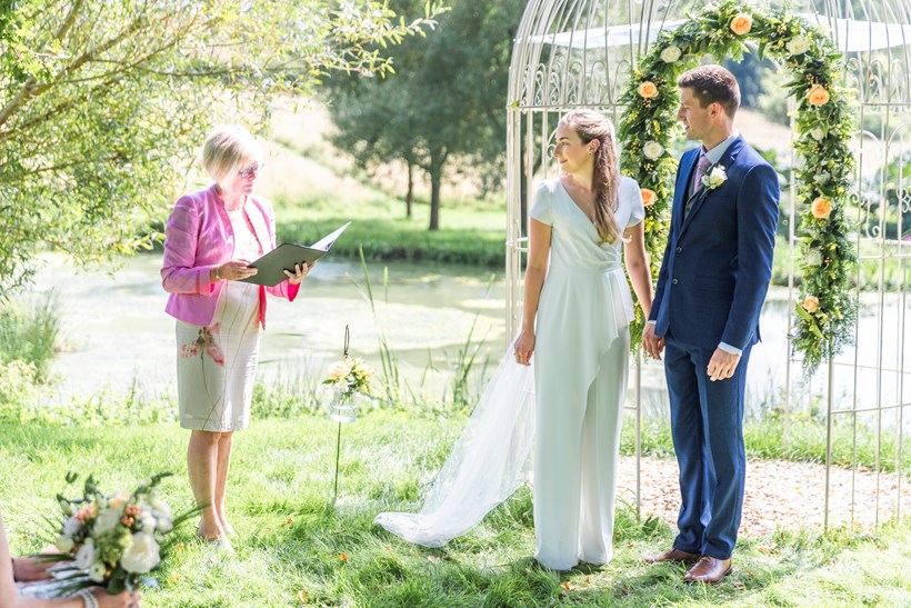 Bride in jumpsuit with groom in front of arch at Ash barton Devon wedding venue