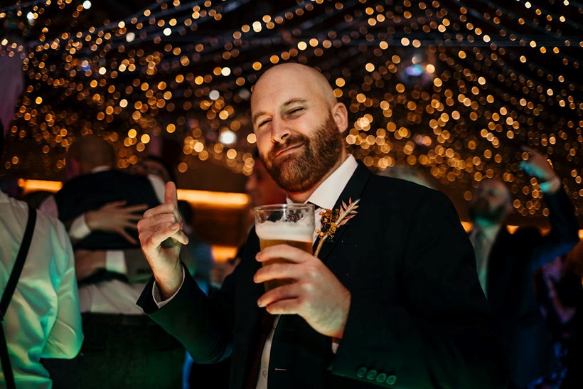 Man with glass of beer with fairy lights in the background at Ash Barton Devon Wedding Venue