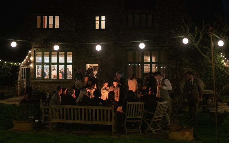 Ashbarton Estate - Wedding guests gather round the fire pit at night at Ash Barton wedding venue Devon